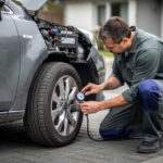 Comment entretenir sa voiture quand elle roule peu toute l’année Voiture citadine garée dans une allée : un homme vérifie la batterie et la pression des pneus.
