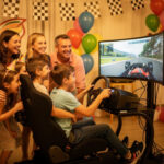 Comment organiser un anniversaire sur le thème du pilotage automobile ? Des enfants et des adultes souriants réunis autour d’un simulateur de conduite dans un décor de circuit automobile, avec des drapeaux à damier, des ballons colorés et une ambiance d’anniversaire chaleureuse.